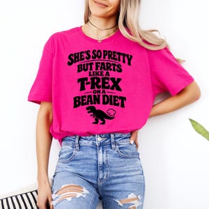 Peut inclure: T-shirt rose vif avec l'inscription noire : "She's so pretty but farts like a T-Rex on a bean diet". Un dessin de dinosaure noir est en dessous. Le t-shirt est porté avec un jean bleu.