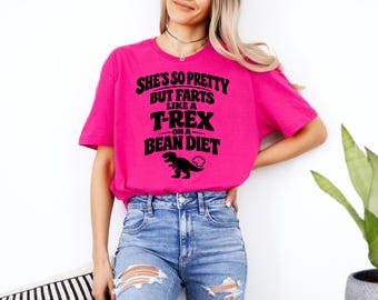 May include: A bright pink t-shirt with the text "She's so pretty but farts like a T-Rex on a bean diet" in black. A black dinosaur graphic is below the text. The shirt is worn with blue jeans.