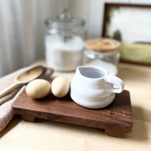 May include: A white ceramic creamer with a handle sits on a wooden cutting board with two eggs. The cutting board has the words "Glad Sparrow Handmade Goods" engraved on it.