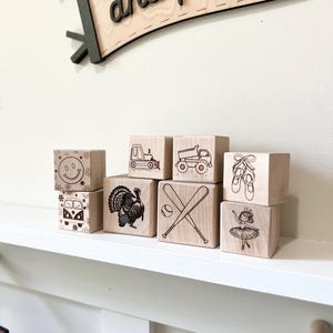 May include: Wooden blocks with various images, including a smiley face, a van, a turkey, baseball bats and a ball, a dump truck, a construction vehicle, ballet shoes, and a ballerina. The blocks are arranged on a white shelf with a wooden sign above that says "go out and play!"