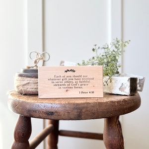 May include: A wooden sign with the text "Each of you should use whatever gift you have received to serve others, as faithful stewards of God's grace in various forms. 1 Peter 4:10." The sign is on a wooden stool with a spool of twine and greenery.