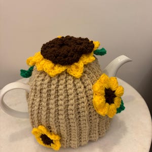 Puede incluir: Una tetera de cerámica blanca cubierta por una funda de ganchillo de color beige. La funda presenta girasoles de ganchillo tridimensionales con centros de color marrón oscuro y pétalos amarillos brillantes, junto con pequeñas hojas verdes.