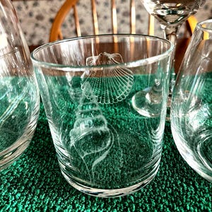May include: Clear drinking glasses with etched seashell designs. The glasses are cylindrical and feature detailed illustrations of various seashells. The image shows multiple glasses, suggesting a set, with a focus on the intricate artwork.