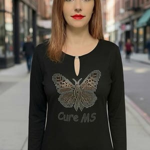 Puede incluir: Camiseta negra de manga larga con un diseño de mariposa de pedrería y la inscripción "Cure MS". La camiseta tiene un escote de ojo de cerradura. La camiseta la lleva una persona.