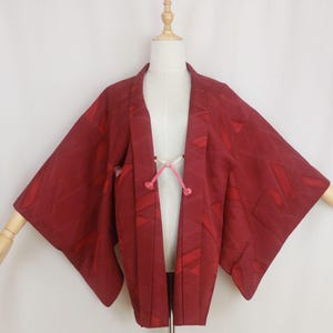 Op de afbeelding: Bordeaux kimono-achtige jas met een geometrisch patroon. De jas wordt getoond op een mannequin met houten armen. Een roze koord sluit de jas aan de voorkant. De stof heeft een subtiele glans.