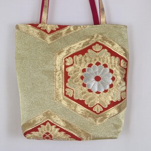 Puede incluir: Un bolso tote de tela dorada metalizada y roja con un patrón hexagonal repetitivo. El diseño incluye motivos florales bordados en hilo plateado y dorado. El bolso tiene dos asas finas de tela roja unidas al borde superior.