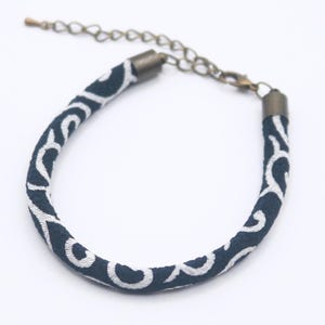 May include: A navy blue bracelet with a white swirling pattern. The bracelet has a bronze-colored chain and clasp for adjustable sizing. The design is a repeating pattern of white swirls on a dark blue background. The bracelet is a jewelry item.