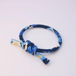 Op de afbeelding: Een marineblauwe armband met een bloemenpatroon in blauw en wit. De armband is geknoopt, met de uiteinden die uitsteken. Het ontwerp is eenvoudig en elegant, geschikt voor dagelijks gebruik. De diameter van de armband is ongeveer 6 cm.