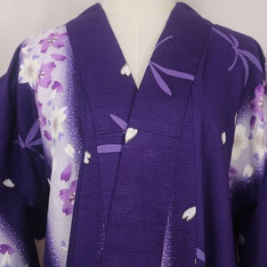 Op de afbeelding: Een dieppaarse kimono met een wit kleurverloop langs de schouders en mouwen. Het kledingstuk is versierd met een print van lichtpaarse en witte kersenbloesems en verspreide bloemblaadjes, uitgevoerd in een textuurrijke stof.