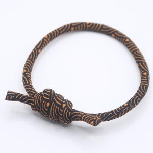 Puede incluir: Una pulsera circular hecha de un cordón fino y flexible con un patrón repetitivo de líneas onduladas negras y naranjas. El cordón está atado en un nudo prominente de varias capas en la parte delantera, con dos extremos cortos que sobresalen.
