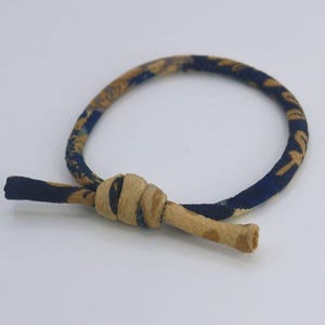 Puede incluir: Una goma para el pelo circular de tela azul oscuro con un estampado floral beige. La banda está asegurada con un nudo central de tela beige lisa, con dos extremos cortos y sueltos que sobresalen del nudo para un acabado decorativo.
