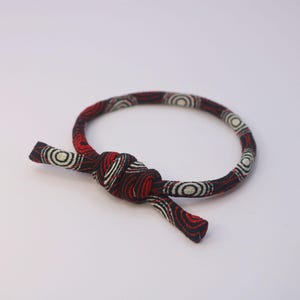 Op de afbeelding: Een armband met rood, zwart en wit patroon, met een geknoopte sluiting. De armband heeft een herhalend cirkelvormig ontwerp in wit en zwart tegen een rode achtergrond. De uiteinden van de armband zijn met elkaar geknoopt.