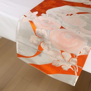 May include: A rectangular table runner featuring a vibrant orange background with a repeating hexagonal pattern. The runner is decorated with large, pale pink floral motifs and flowing silver-grey ribbons that drape across the fabric surface.