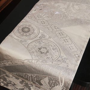 以下が含まれることがあります： ベージュとグレーの装飾的なテーブルランナー。複雑な模様で、円形と幾何学的なデザイン、鳥の画像が特徴です。生地は微妙な光沢があります。テーブルランナーは黒いテーブルの上に置かれています。
