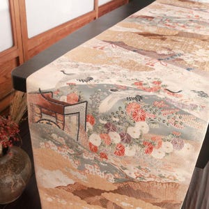 May include: A decorative table runner with a Japanese-inspired design. The runner features a landscape with cranes, flowers, and a traditional building in shades of beige, blue, and red. The runner is placed on a dark surface.