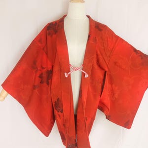 以下が含まれることがあります： 全体に同系色の花柄があしらわれた鮮やかな赤色の羽織。袖は幅広でゆったりとしており、前合わせは白と赤の細い組み紐で結ばれており、構造的で開いたフロントスタイルを作り出しています。