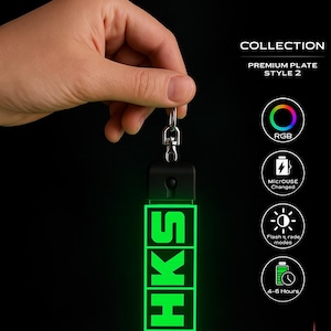 Pode incluir: Um porta-chaves preto com as letras "HKS" iluminadas em verde. O porta-chaves tem uma corrente prateada e uma parte superior preta. O texto adicional inclui "COLLECTION" e "PREMIUM PLATE STYLE 2".