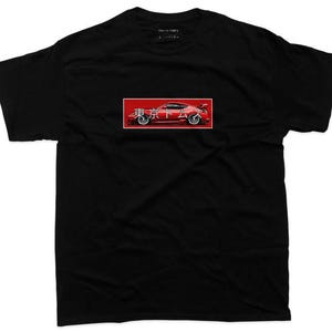 May include: Black t-shirt featuring a red rectangle with a car graphic and Japanese text. The car is red with white accents. The shirt has a crew neck and short sleeves. The brand name "TOKYO TOM'S" is printed on the top.