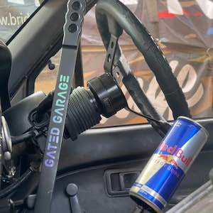 Peut inclure: Int&eacute;rieur de voiture avec un volant noir, un levier de vitesse noir avec le texte "GATED GARAGE" et une canette de boisson &eacute;nergisante Red Bull. Le levier de vitesse comporte une s&eacute;rie de trous. Le volant est noir et a une poign&eacute;e en cuir.