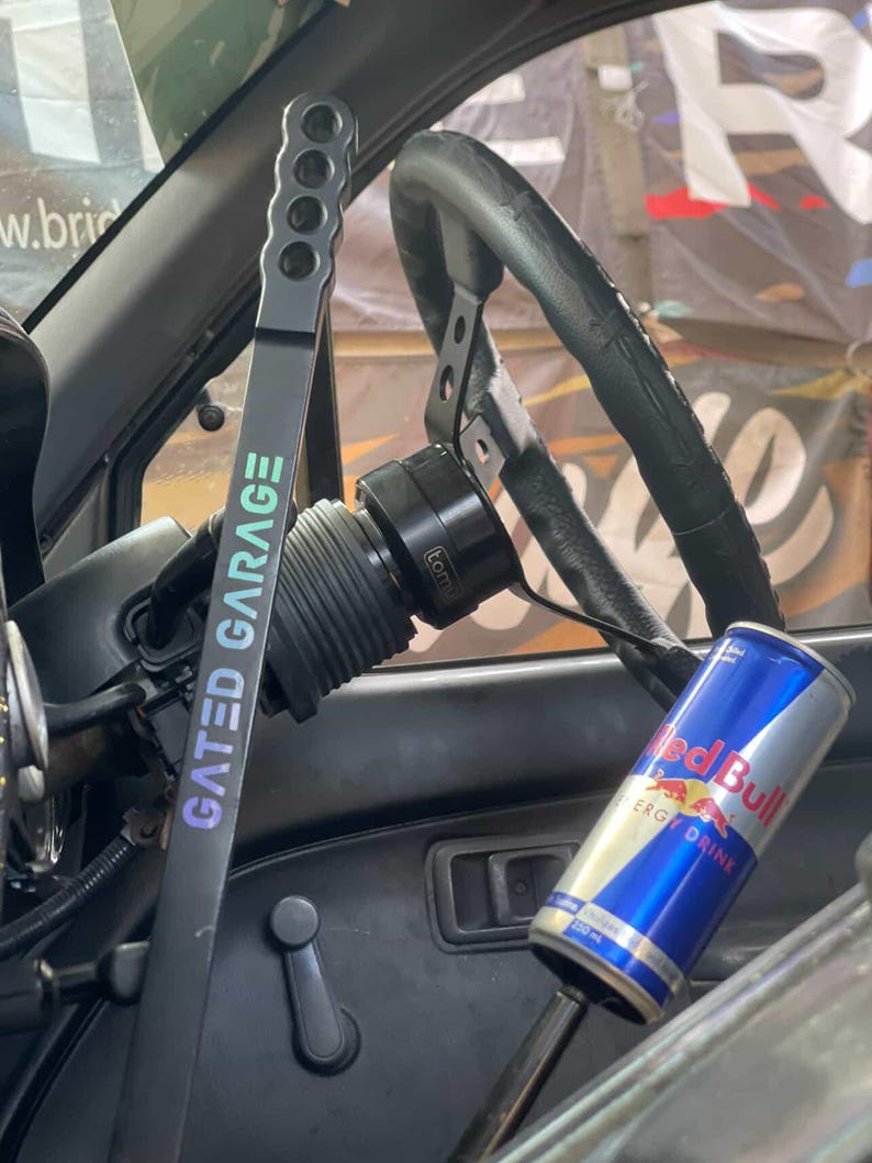 Peut inclure: Int&eacute;rieur de voiture avec un volant noir, un levier de vitesse noir avec le texte "GATED GARAGE" et une canette de boisson &eacute;nergisante Red Bull. Le levier de vitesse comporte une s&eacute;rie de trous. Le volant est noir et a une poign&eacute;e en cuir.