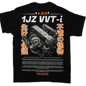 Op de afbeelding: Zwart T-shirt met een witte afbeelding van een 1JZ VVT-i-motor met de tekst "1JZ VVT-i", "トヨタ", "生ける伝説" en "不滅の怪物" in het Japans en oranje tekst.