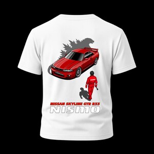 Puede incluir: Camiseta blanca con un gráfico de un coche rojo Nissan Skyline GTR R33, una silueta de Godzilla y una figura con un traje de carreras rojo. El texto "NISSAN SKYLINE GTR R33 NISMO" está impreso debajo.