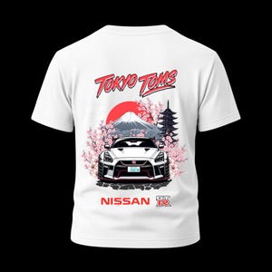 May include: White t-shirt featuring a graphic of a white sports car with a red accent, set against a backdrop of Mount Fuji, cherry blossoms, and a pagoda. The text "Tokyo Toms" and "Nissan GT-R" are also displayed.