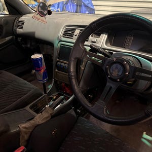 Peut inclure: Int&eacute;rieur de voiture avec volant, tableau de bord et si&egrave;ges noirs. Une canette de Red Bull est fix&eacute;e au levier de vitesse. Un d&eacute;sodorisant noir avec le texte "TOKYO TON 5" est suspendu au tableau de bord.
