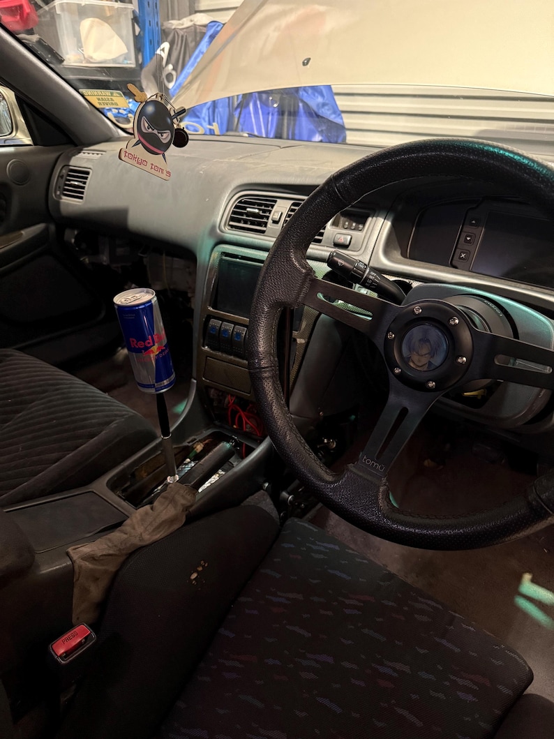 Peut inclure: Int&eacute;rieur de voiture avec volant, tableau de bord et si&egrave;ges noirs. Une canette de Red Bull est fix&eacute;e au levier de vitesse. Un d&eacute;sodorisant noir avec le texte "TOKYO TON 5" est suspendu au tableau de bord.