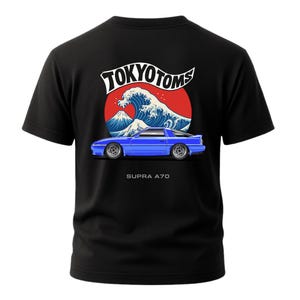 May include: Black t-shirt featuring a blue sports car graphic with the text "SUPRA A70" below it. The design includes a red circle with a white wave and mountain scene, and the words "TOKYO TOMS" in a stylized font.