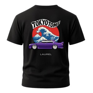 May include: Black t-shirt featuring a graphic of a purple car with white wheels, set against a red circle with a wave and mountain design. The text "TOKYO TOMS" and "LAUREL" are also visible.