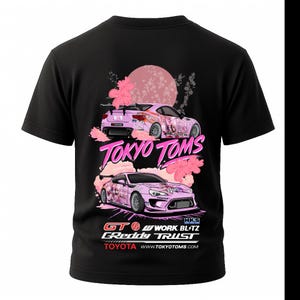 May include: Black t-shirt featuring two pink race cars with floral accents and the text "Tokyo Toms." The design includes a pink sun and Japanese text. Additional text includes "GT," "WORK," "BLITZ," "GReddy TRUST," and "TOYOTA."