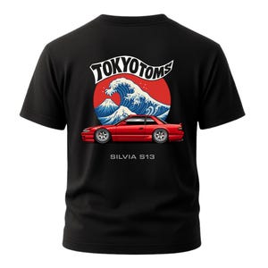 May include: Black t-shirt featuring a red car graphic with the text "SILVIA S13" below it. Above the car is a design of a wave, a mountain, and the text "TOKYO TOMS" in a stylized font. The design is set against a red circle.