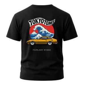 May include: Black t-shirt featuring a graphic of a yellow Fairlady S130Z car with a wave and mountain design, and the text "TOKYO TOMS". The shirt has short sleeves and a crew neck.