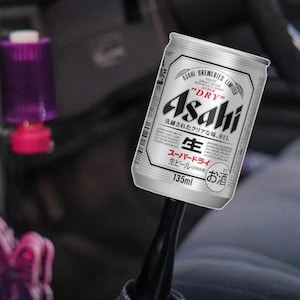May include: A silver Asahi Super Dry beer can, 135ml, with Japanese text and a white and black label. The can is held against a blurred background. The text on the can includes "Asahi Breweries Limited" and "Dry".