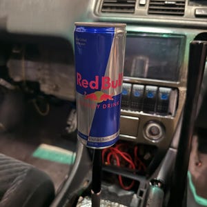 Peut inclure: Une canette de boisson &eacute;nergisante Red Bull fix&eacute;e au levier de vitesse d'une voiture. La canette est bleue et argent&eacute;e avec le logo Red Bull. L'int&eacute;rieur de la voiture est visible en arri&egrave;re-plan.