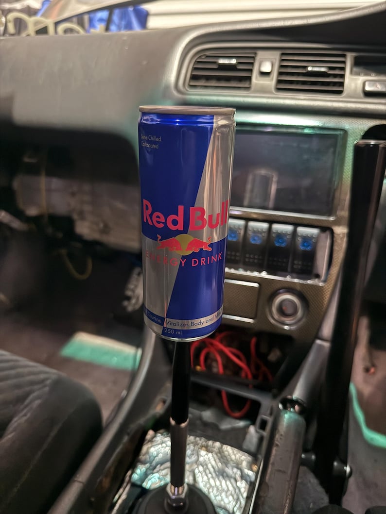 Peut inclure: Une canette de boisson &eacute;nergisante Red Bull fix&eacute;e au levier de vitesse d'une voiture. La canette est bleue et argent&eacute;e avec le logo Red Bull. L'int&eacute;rieur de la voiture est visible en arri&egrave;re-plan.