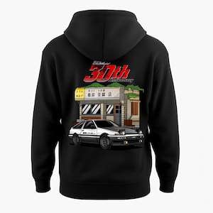 May include: Black hooded sweatshirt with a graphic of a white and black car in front of a Japanese tofu shop. The design includes the text "30th Anniversary" in red and white, and Japanese text.