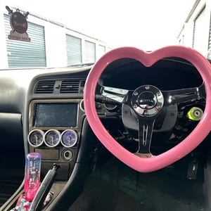 Könnte beinhalten: Autoinnenraum mit einem pinkfarbenen, herzförmigen Lenkrad. Das Armaturenbrett ist schwarz mit Anzeigen und einem kleinen Bildschirm. Ein dekorativer Lufterfrischer hängt am Rückspiegel.