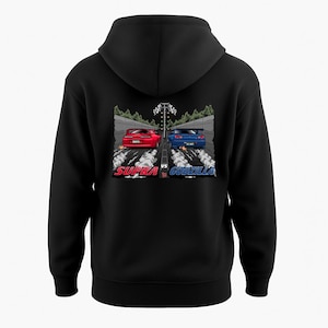 May include: Black hooded sweatshirt featuring a graphic of two cars racing, a red Supra and a blue Godzilla, with the words "Supra vs Godzilla" in red and blue. The design includes a checkered flag and a forest background.