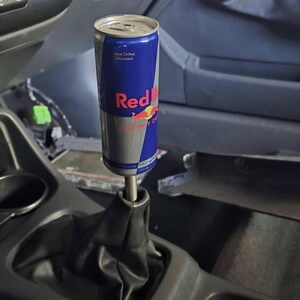 Peut inclure: Un canette de boisson &eacute;nergisante Red Bull modifi&eacute;e en pommeau de levier de vitesse de voiture. La canette, bleue, argent&eacute;e et rouge, porte le logo et le texte Red Bull. Mont&eacute;e sur un soufflet en cuir noir &agrave; l'int&eacute;rieur d'une voiture.