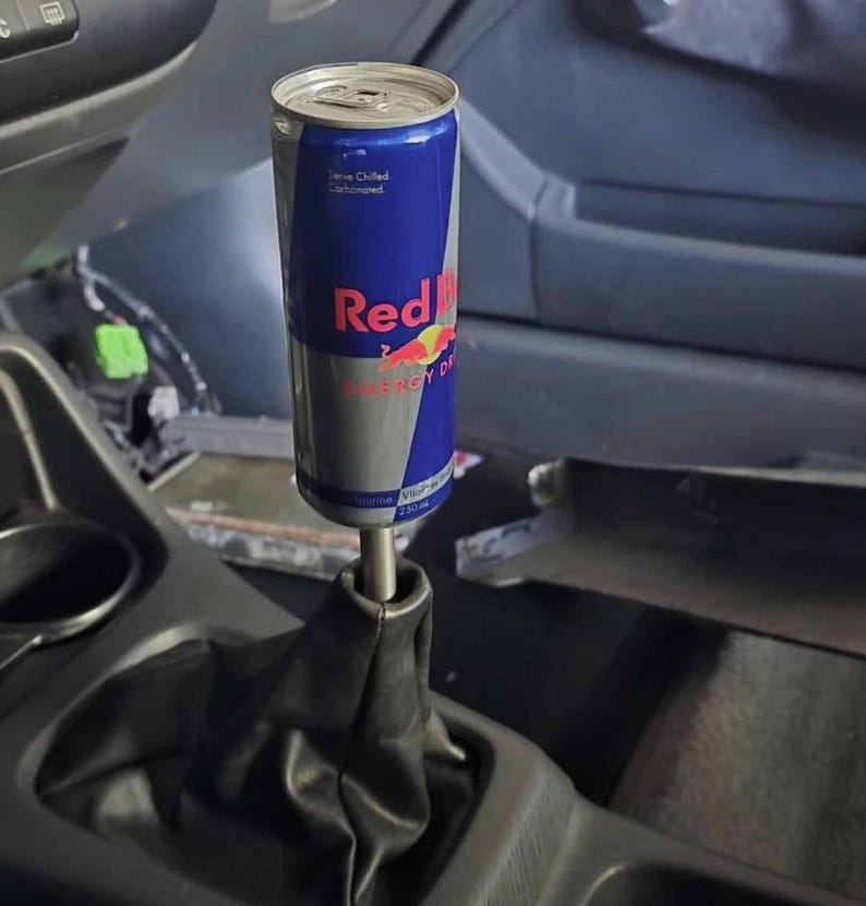 Peut inclure: Un canette de boisson &eacute;nergisante Red Bull modifi&eacute;e en pommeau de levier de vitesse de voiture. La canette, bleue, argent&eacute;e et rouge, porte le logo et le texte Red Bull. Mont&eacute;e sur un soufflet en cuir noir &agrave; l'int&eacute;rieur d'une voiture.