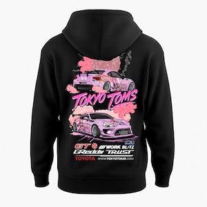 Peut inclure: Sweat à capuche noir avec un motif rose de deux voitures de course, des détails de fleurs de cerisier et le texte "TOKYO TOMS". Le texte supplémentaire comprend "GT", "GReddy TRUST" et "TOYOTA". Le motif est au dos du sweat à capuche.