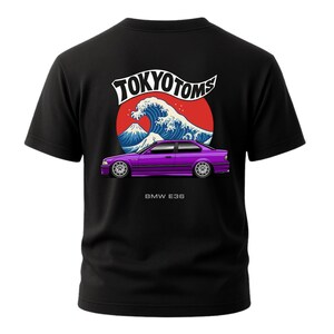Puede incluir: Camiseta negra con un gráfico de coche morado y el texto "BMW E36". El diseño incluye un círculo rojo con una ola blanca y una escena de montaña, y el texto "TOKYO TOMS".