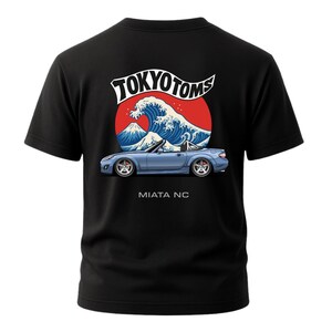 May include: Black t-shirt featuring a graphic of a blue convertible car with the text "MIATA NC" below it. Above the car is a red circle with a white wave and mountain design, and the text "TOKYO TOMS".
