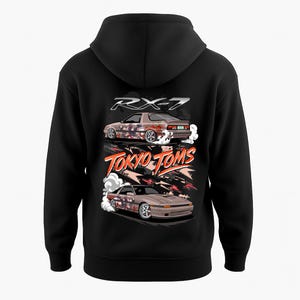 Op de afbeelding: Zwarte hoodie met een afbeelding van twee bruine RX-7 auto's met rooksporen. De tekst "RX-7" staat boven de auto's en "TOKYO TOMS" eronder in oranje en wit. Het ontwerp staat op de achterkant van de hoodie.
