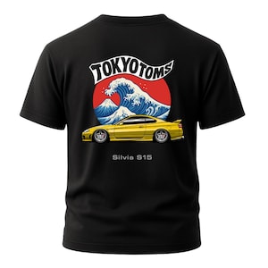 May include: Black t-shirt featuring a yellow sports car design with the text "Silvia S15." The graphic includes a red sun, a blue wave, and the words "TOKYO TOMS" in white. The shirt is short-sleeved.