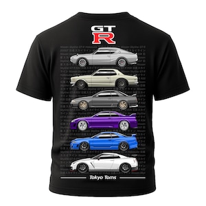 Puede incluir: Camiseta negra con un gráfico de cinco coches Nissan Skyline GT-R de varios colores. Los coches están apilados verticalmente, con el texto "Tokyo Toms" en la parte inferior. El logotipo GT-R está en la parte superior.