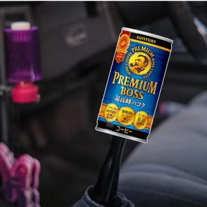 May include: A can of Suntory Premium Boss Coffee, with a blue and gold label and Japanese text. The can displays the words "Premium Boss" and "Coffee". The can is held in a vehicle.