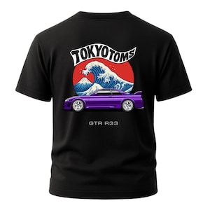 May include: Black t-shirt featuring a purple sports car graphic with the text "GTR R33" below it. Above the car is a design of a wave, a mountain, and the text "TOKYO TOMS" in white.